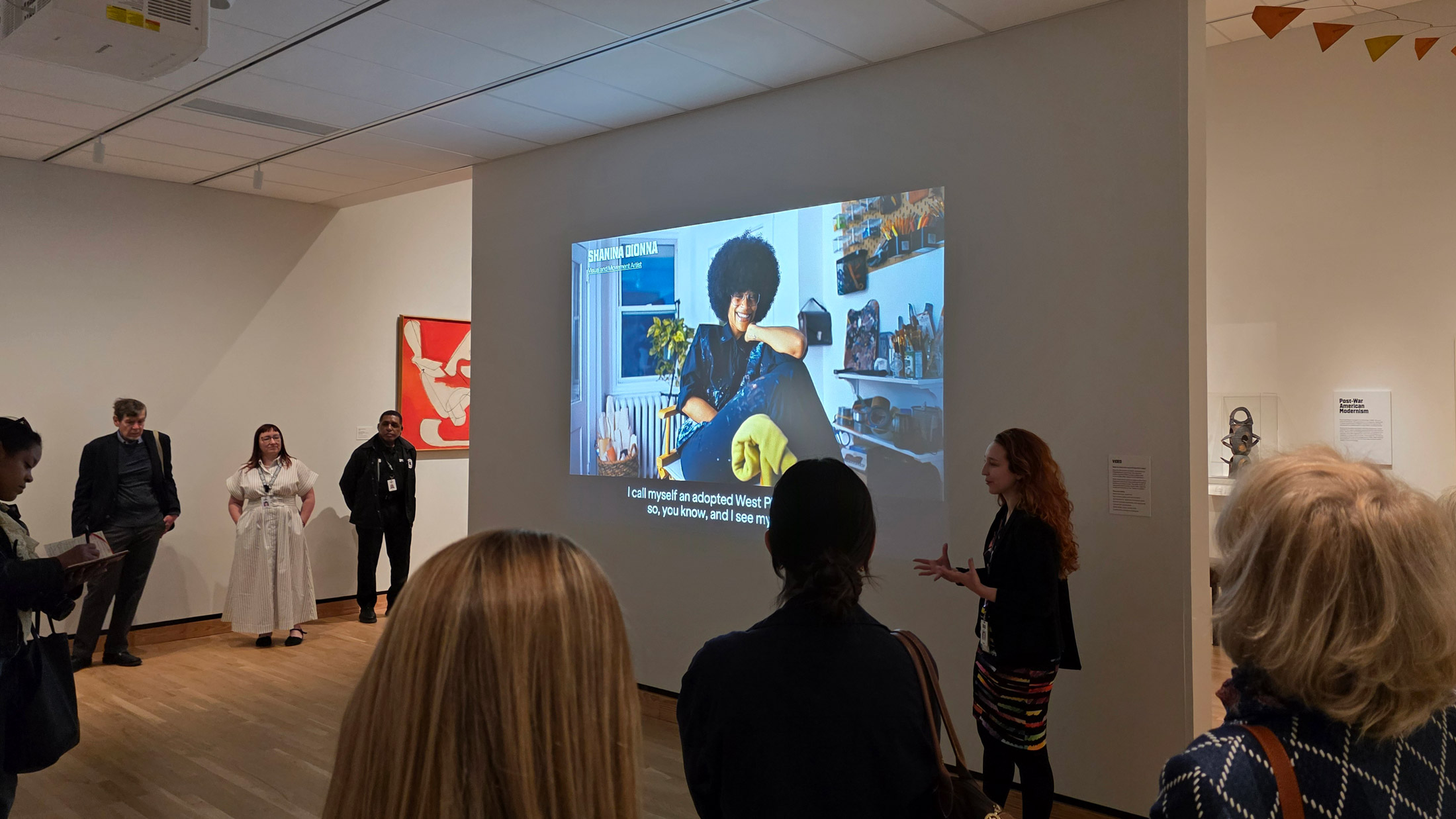Visitors gather in a gallery during Philadelphia’s A Nation of Artists exhibit as a presenter speaks beside a wall projection showing a video interview with an artist, with additional artworks displayed around the room.