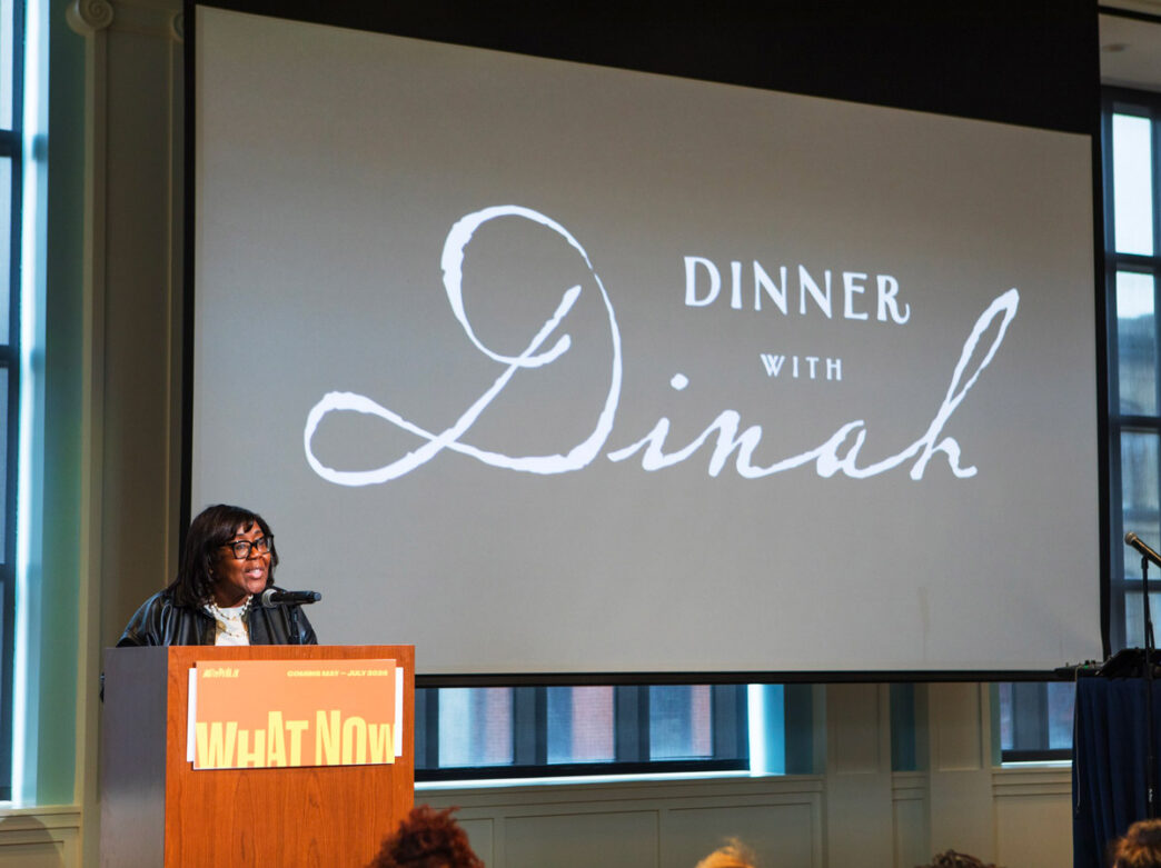 Speaker presenting at the ArtPhilly "What Now" program about "Dinner with Dinah" event at a podium with projection screen.