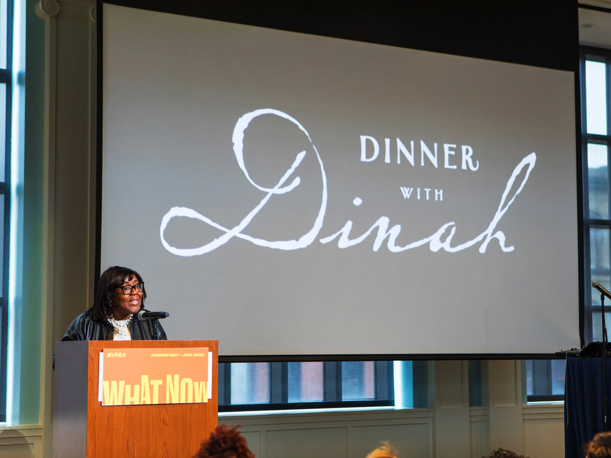 Speaker presenting at the ArtPhilly "What Now" program about "Dinner with Dinah" event at a podium with projection screen.