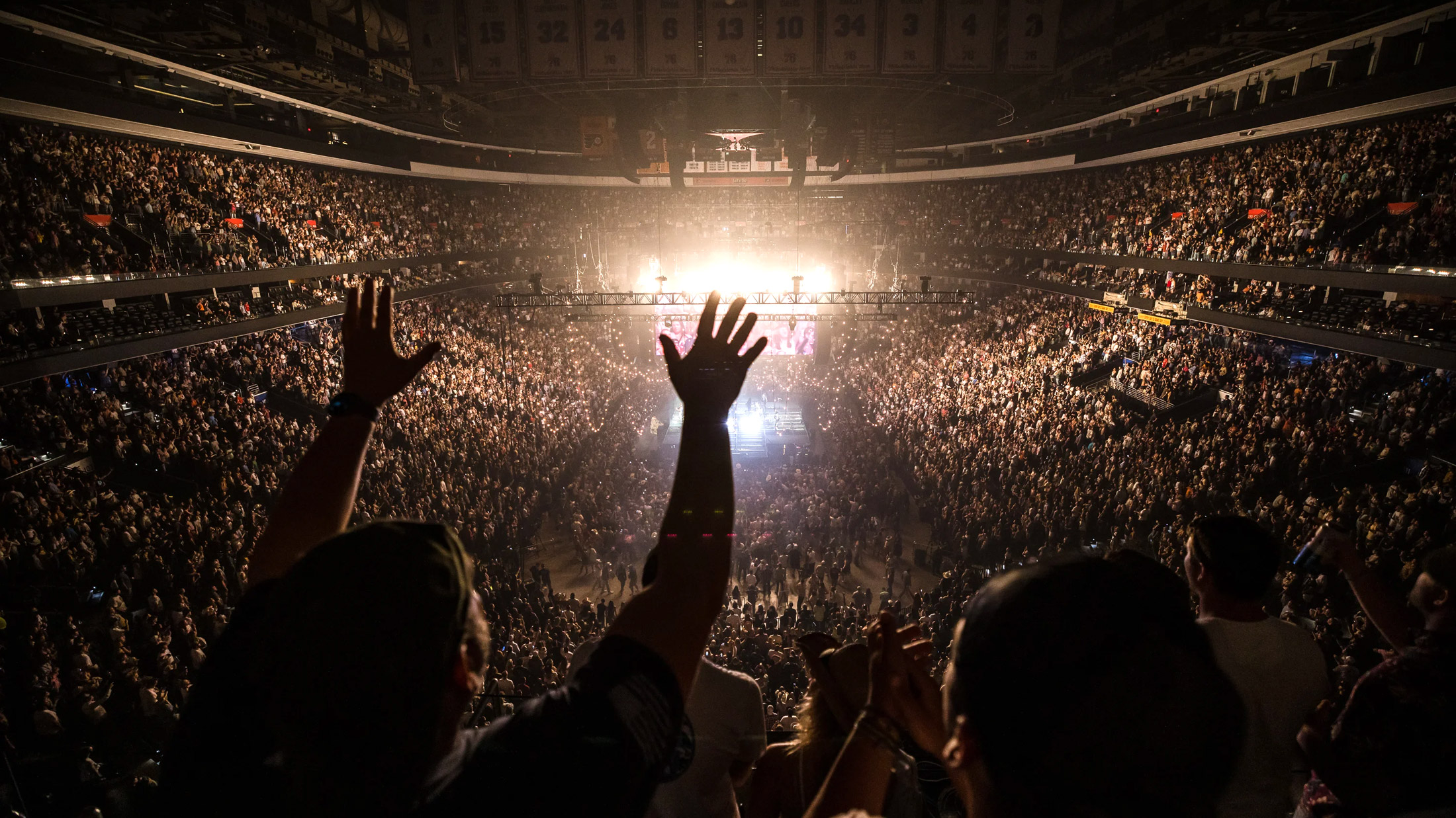 Crowd fills Xfinity Mobile Arena in Philadelphia during a concert, with thousands of fans cheering and raising their hands toward a brightly lit stage at center.