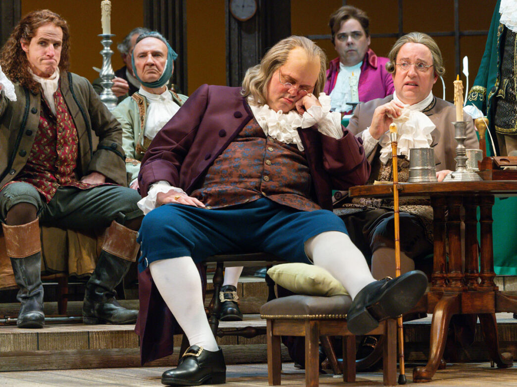 Een scène uit "1776: The Musical" toont vijf acteurs in 18e-eeuwse koloniale kostuums op het podium. Een centrale figuur in een bordeauxrode jas en blauwe broek zit prominent met zijn hand voor zijn gezicht en lijkt te slapen. Vier andere mannen in historische kleding staan en zitten om hem heen in een formeel interieur met kandelaars, een vlag en houten meubilair, dat een moment uitbeeldt tijdens de Amerikaanse Revolutie.