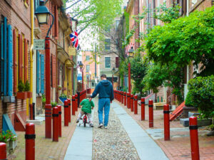 A father walks aside his son, who rides a tricycle, on the brick-lined street of Elfreth's Alley with historic brick buildings on either side of the narrow road.