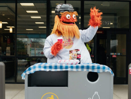 Gritty, Philadelphia Flyers mascot, holds a cup of coffee while waving. A tablecloth is draped over a trashcan in front of him, which is topped with a hoagie, chips and soda.