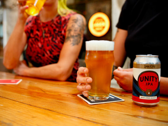 A man and a woman sit at a bar. The man holds a pint full of beer while the woman takes a sip of hers.