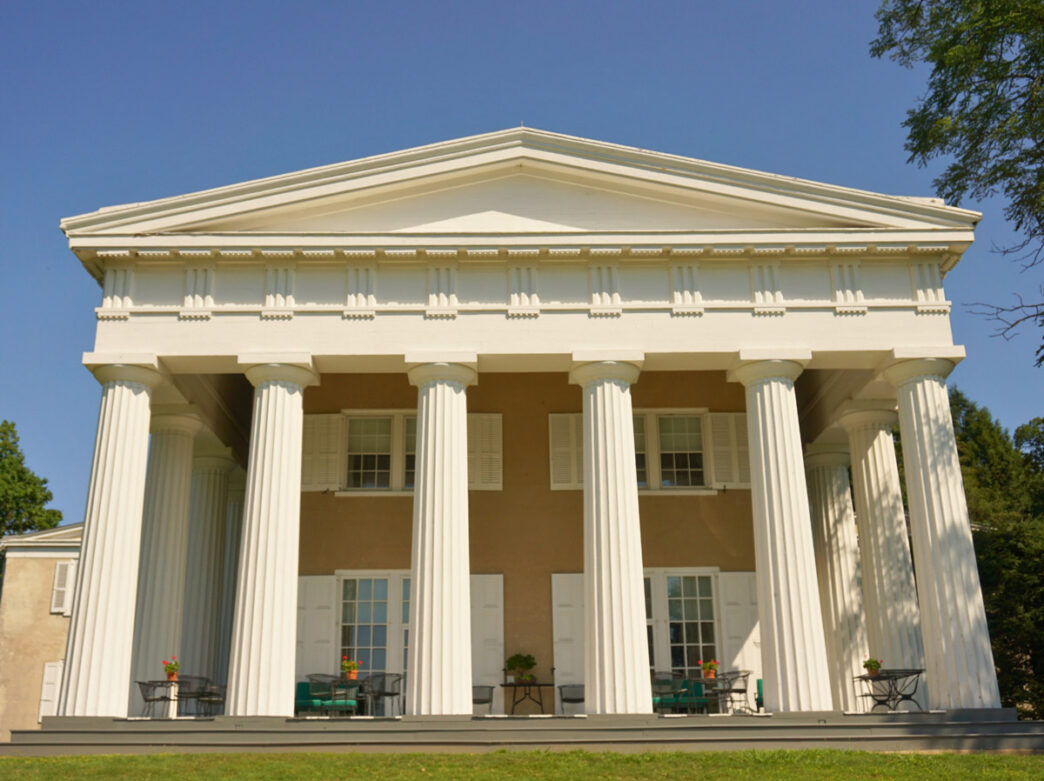 Front view of the historic Greek Revival mansion at Andalusia Historic House, Gardens & Arboretum, featuring towering white columns, a triangular pediment, and a broad porch set against a bright blue sky.