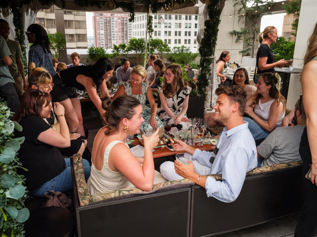 A lively rooftop gathering at Attico in Philadelphia, with groups of young adults socializing around low tables under a pergola wrapped in greenery and string lights. Guests are seated and standing with cocktails and shared plates, while the surrounding city skyline and neighboring high-rise buildings create an upscale urban backdrop.