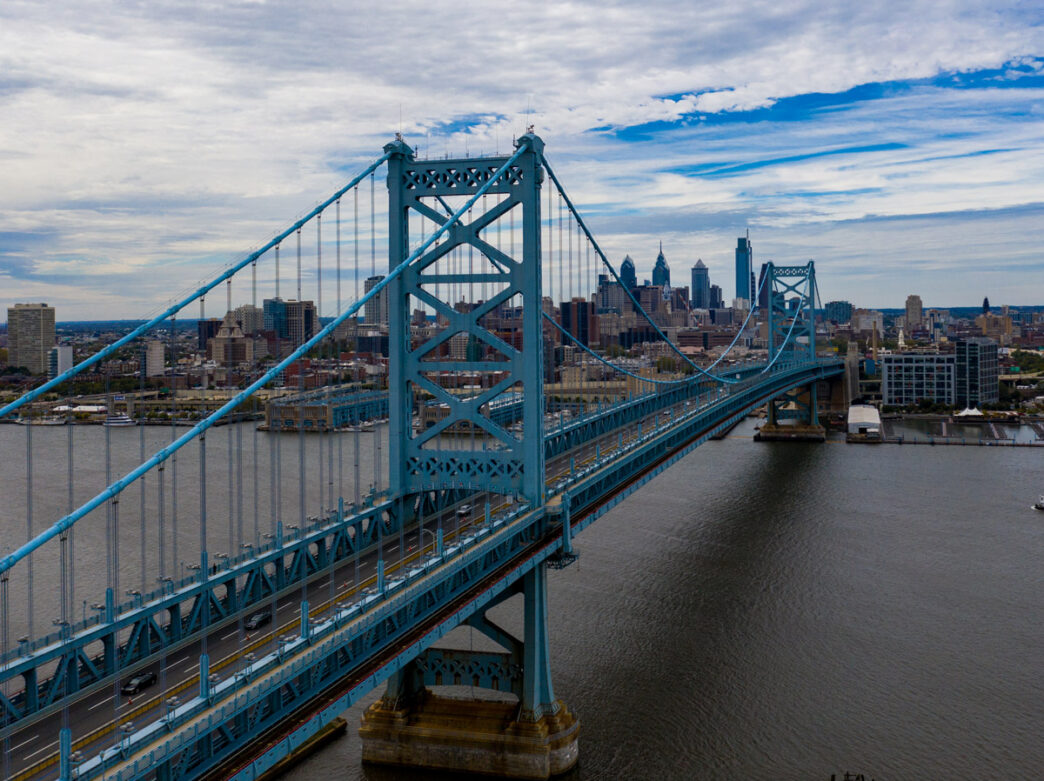 The iconic teal-colored Benjamin Franklin Bridge spans the Delaware River, connecting Philadelphia to Camden, New Jersey. The suspension bridge's steel cables and X-braced towers are prominently featured, with the Philadelphia skyline and City Hall visible in the background against a cloudy blue sky.