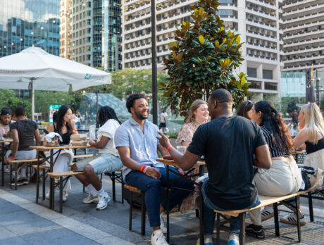 Een groep mensen geniet van een drankje en socialiseert aan buitentafels in Dilworth Park in het centrum van Philadelphia, met hoge wolkenkrabbers en bomen zichtbaar op de achtergrond.