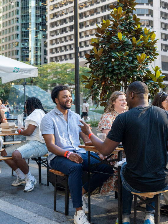 A group of people enjoy drinks and socialize at outdoor tables in Dilworth Park in downtown Philadelphia, with tall skyscrapers and trees visible in the background.
