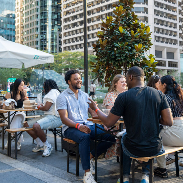 A group of people enjoy drinks and socialize at outdoor tables in Dilworth Park in downtown Philadelphia, with tall skyscrapers and trees visible in the background.