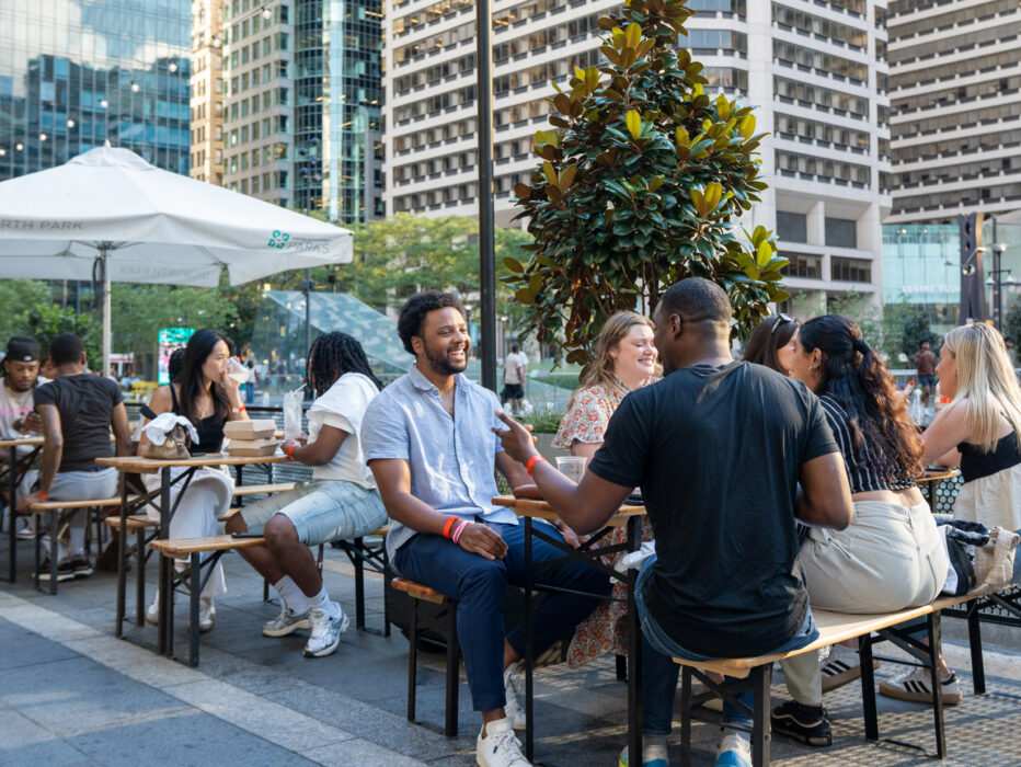 Een groep mensen geniet van een drankje en socialiseert aan buitentafels in Dilworth Park in het centrum van Philadelphia, met hoge wolkenkrabbers en bomen zichtbaar op de achtergrond.