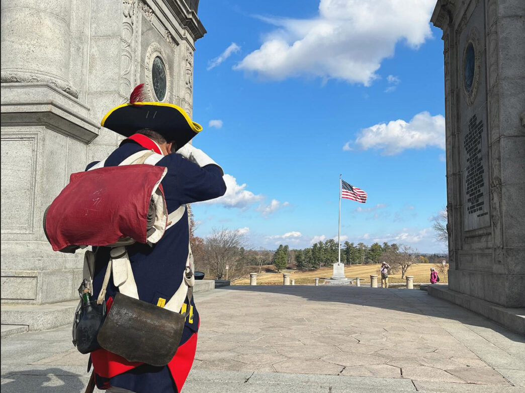 A person dressed in a colonial Revolutionary War-era military uniform stands between two stone buildings at Valley Forge National Historical Park, looking toward an American flag flying on a flagpole in the distance across an open field.