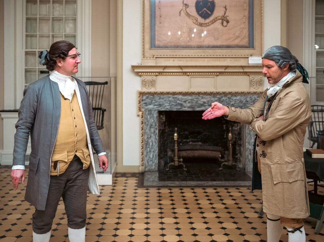 Two historical reenactors in 18th-century colonial clothing converse inside a historic Philadelphia room with a fireplace, checkered floor, and period furnishings.