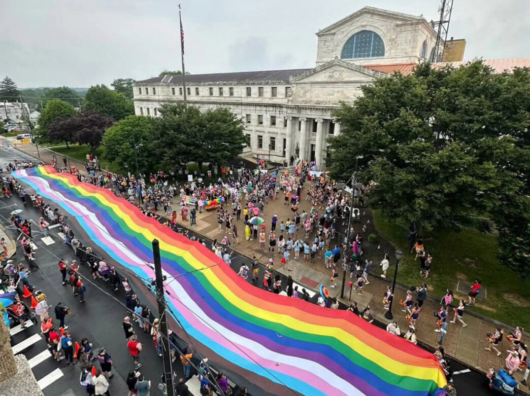 A large rainbow pride flag stretches down a street during Delco Pride in Delaware County, Pennsylvania, as crowds gather outside a historic courthouse building on an overcast day. The aerial view shows people lining the sidewalks and participating in the Pride celebration.