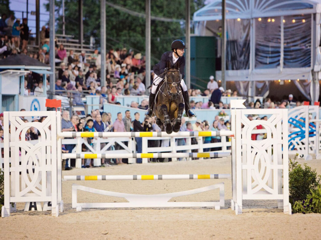 Een ruiter en paard springen over een hindernis tijdens de Devon Horse Show, met toeschouwers op de tribunes rond de arena.