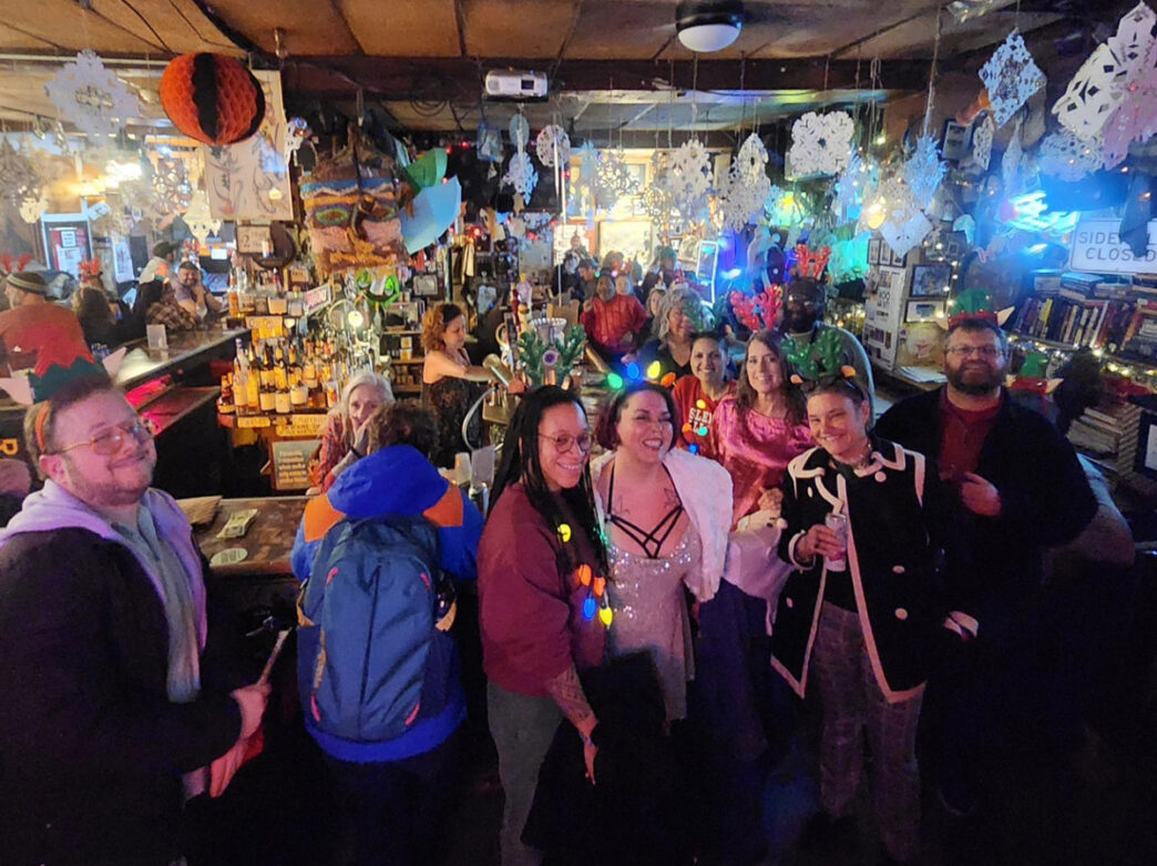 Une foule festive se rassemble à l'intérieur du Dirty Frank's à Philadelphie, avec des clients portant des pulls de Noël, des bois de renne et des colliers lumineux colorés sous des flocons de neige en papier suspendus et un décor de bar éclectique.