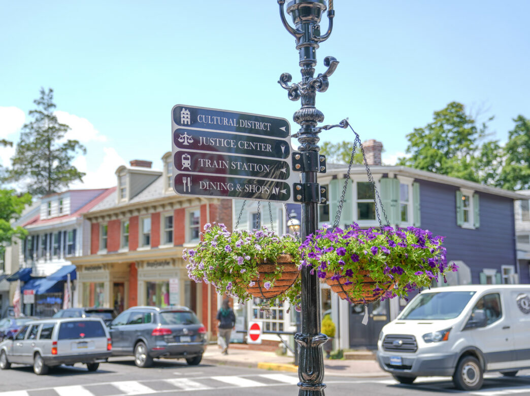Un poste de señalización decorativo en el centro de Doylestown muestra letreros indicativos hacia el Distrito Cultural, el Centro de Justicia, la estación de tren y la zona de restaurantes y tiendas. El poste está adornado con macetas colgantes con petunias moradas y follaje verde. Edificios históricos de ladrillo en tonos rojos, azules y crema bordean la calle arbolada, y se pueden ver vehículos estacionados y peatones a lo largo de la encantadora intersección del centro.