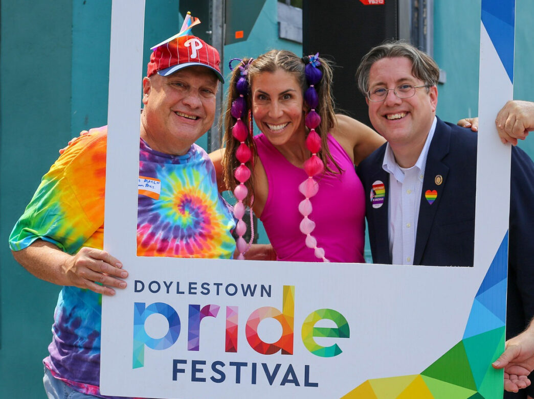 Three people smile and pose inside a large “Doylestown Pride Festival” photo frame at an outdoor Pride celebration in Doylestown, Pennsylvania. One person wears a rainbow tie-dye shirt and Phillies cap, another wears a bright pink outfit with colorful braided hair extensions, and the third wears a suit jacket with rainbow pride pins.