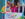 Three people smile and pose inside a large “Doylestown Pride Festival” photo frame at an outdoor Pride celebration in Doylestown, Pennsylvania. One person wears a rainbow tie-dye shirt and Phillies cap, another wears a bright pink outfit with colorful braided hair extensions, and the third wears a suit jacket with rainbow pride pins.