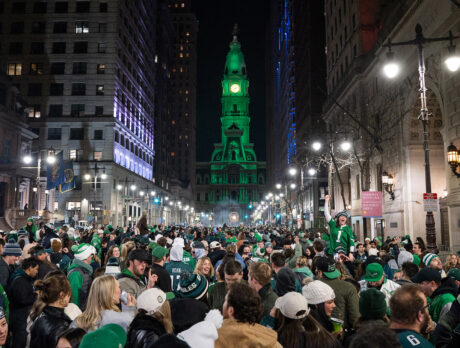 A large crowd of Eagles fans gather in front of City Hall in Philadelphia to celebrate a championship win. The City Hall building is lit up in green.
