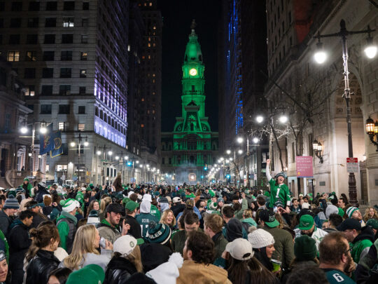 Uma grande multidão de torcedores do Eagles se reúne em frente ao City Hall na Filadélfia para comemorar uma vitória no campeonato. O edifício City Hall está iluminado em verde.