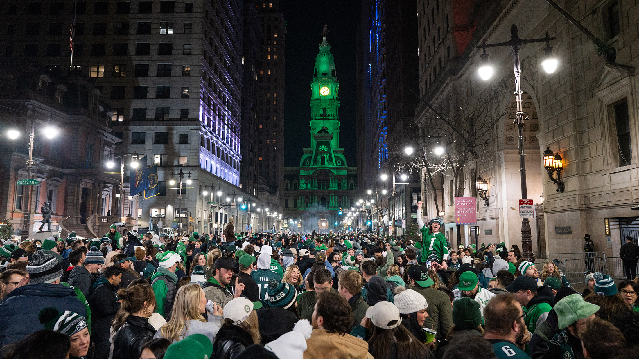 A large crowd of Eagles fans gather in front of City Hall in Philadelphia to celebrate a championship win. The City Hall building is lit up in green.