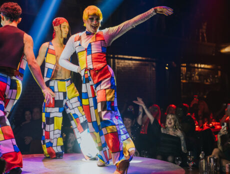 Artistas vestidos con trajes de colores vivos y motivos geométricos bailan en un escenario circular en el Fabrika de Filadelfia, iluminados por espectaculares focos, mientras el público observa y aplaude desde las mesas que rodean el escenario.