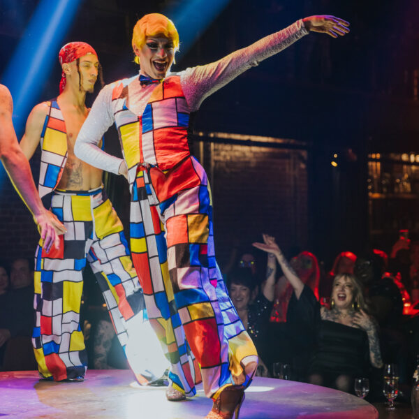 Artistas vestidos con trajes de colores vivos y motivos geométricos bailan en un escenario circular en el Fabrika de Filadelfia, iluminados por espectaculares focos, mientras el público observa y aplaude desde las mesas que rodean el escenario.