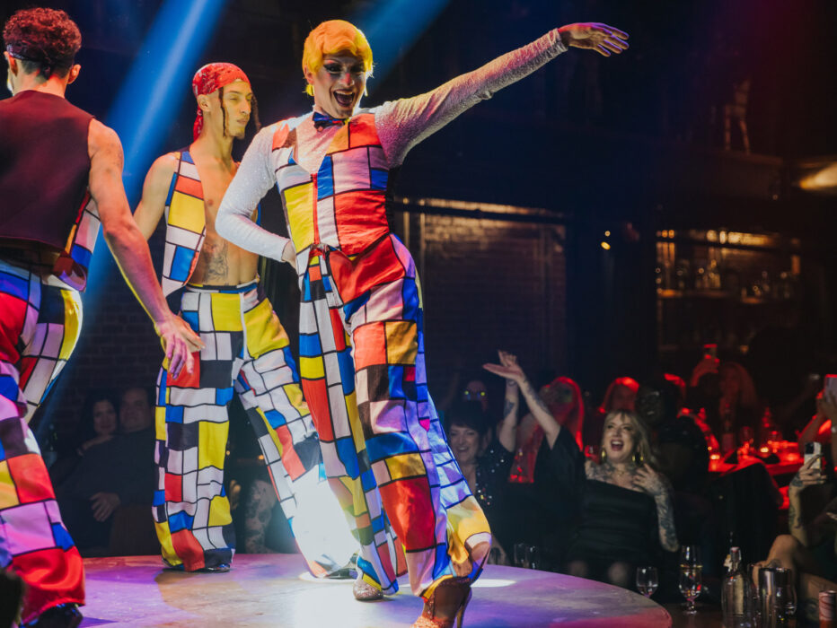 Artistas vestidos con trajes de colores vivos y motivos geométricos bailan en un escenario circular en el Fabrika de Filadelfia, iluminados por espectaculares focos, mientras el público observa y aplaude desde las mesas que rodean el escenario.