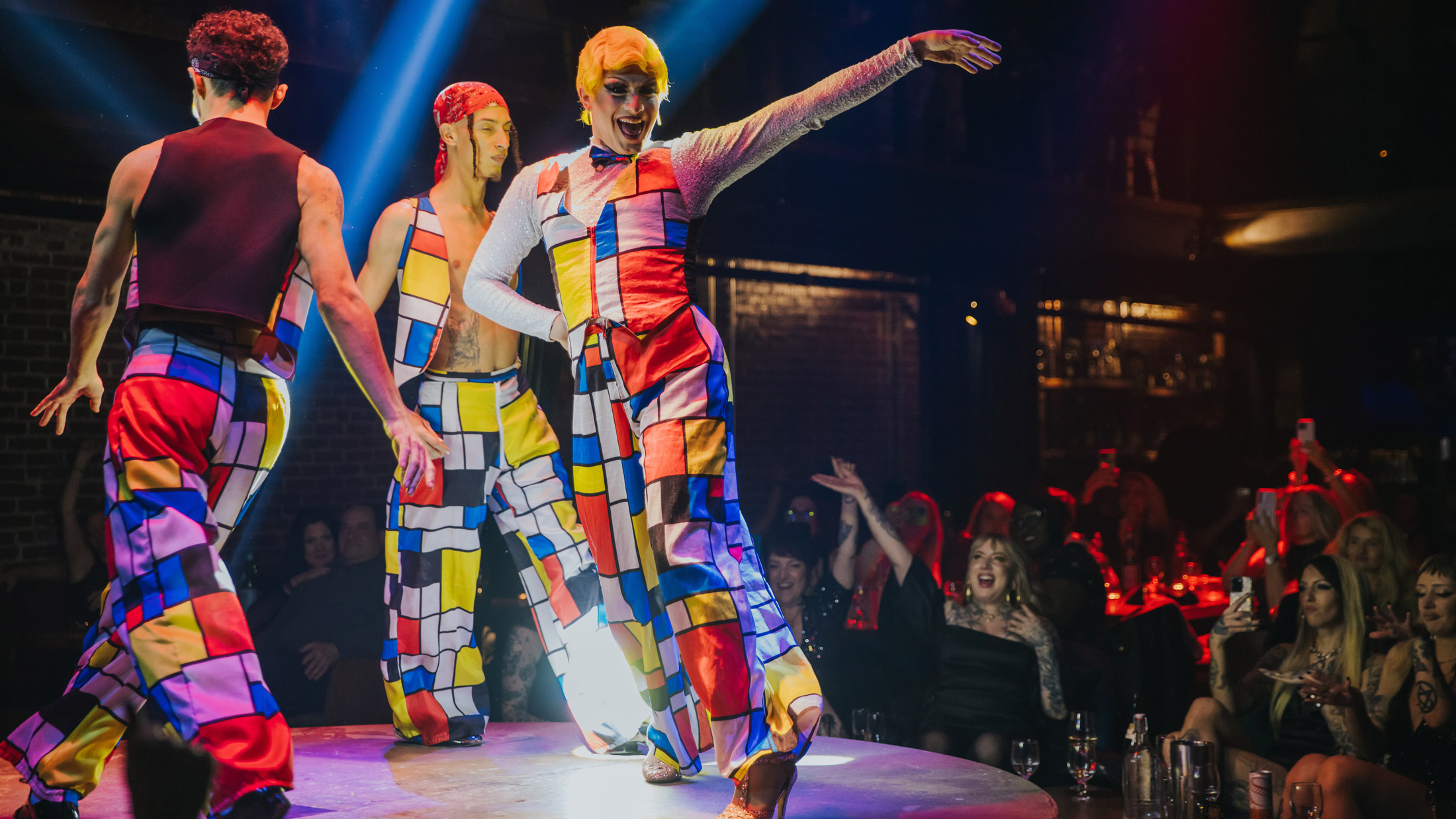 Performers in felgekleurde kostuums met geometrische patronen dansen op een cirkelvormig podium in Fabrika in Philadelphia, verlicht door dramatische spots terwijl het publiek toekijkt en juicht vanaf de omringende tafels.