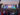 Large crowd gathered in front of a colorful stage at the FIFA World Cup 2026 Fan Festival in Philadelphia, with fans holding up phones and flags as a soccer match plays on a giant central screen.
