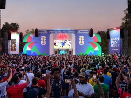 Large crowd gathered in front of a colorful stage at the FIFA World Cup 2026 Fan Festival in Philadelphia, with fans holding up phones and flags as a soccer match plays on a giant central screen.