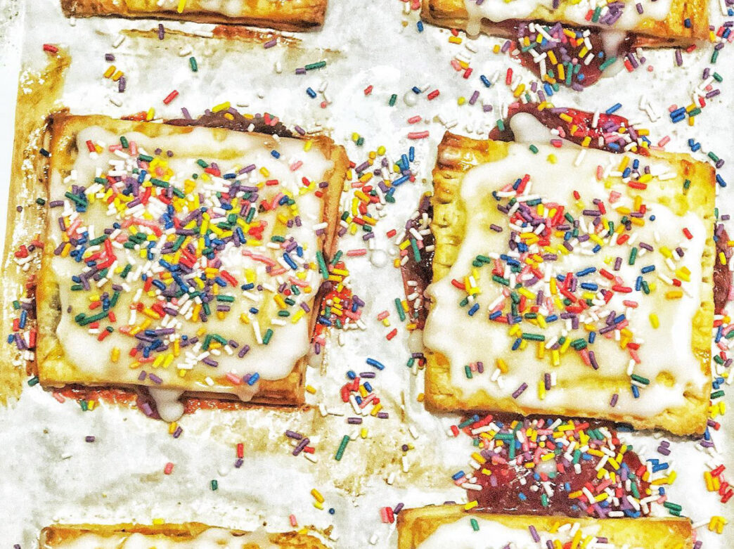 Close-up of gluten-free Pop-Tarts style pastries with golden-brown crusts topped with white icing and rainbow-colored sprinkles, arranged on a white surface.