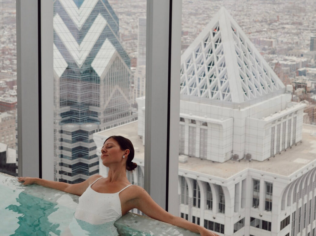 Woman relaxing in an indoor pool at the Four Seasons Philadelphia at Comcast Center spa, with views of iconic downtown skyscrapers through floor-to-ceiling windows.
