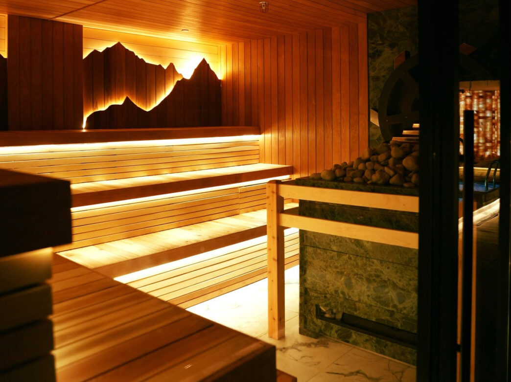 A modern sauna room at Fusion Spa with tiered wooden benches illuminated by warm ambient lighting. A scenic mountain range silhouette is backlit on the wall, and heated stones are visible in a basin, creating a serene wellness atmosphere.