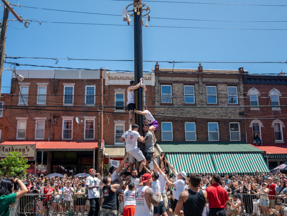 Los participantes trepan por un poste engrasado durante el Festival Italian Market en la calle South 9th Street de Filadelfia. Varios hombres intentan escalar el alto poste eléctrico mientras una gran multitud observa desde detrás de las barreras. Al fondo, la calle está flanqueada por edificios históricos de ladrillo, entre los que se ven los toldos de las tiendas, incluido el de Anthony's. El cielo está despejado y azul, y los participantes, vestidos con ropa deportiva informal, se turnan para trepar por el poste en esta tradicional competencia del festival.