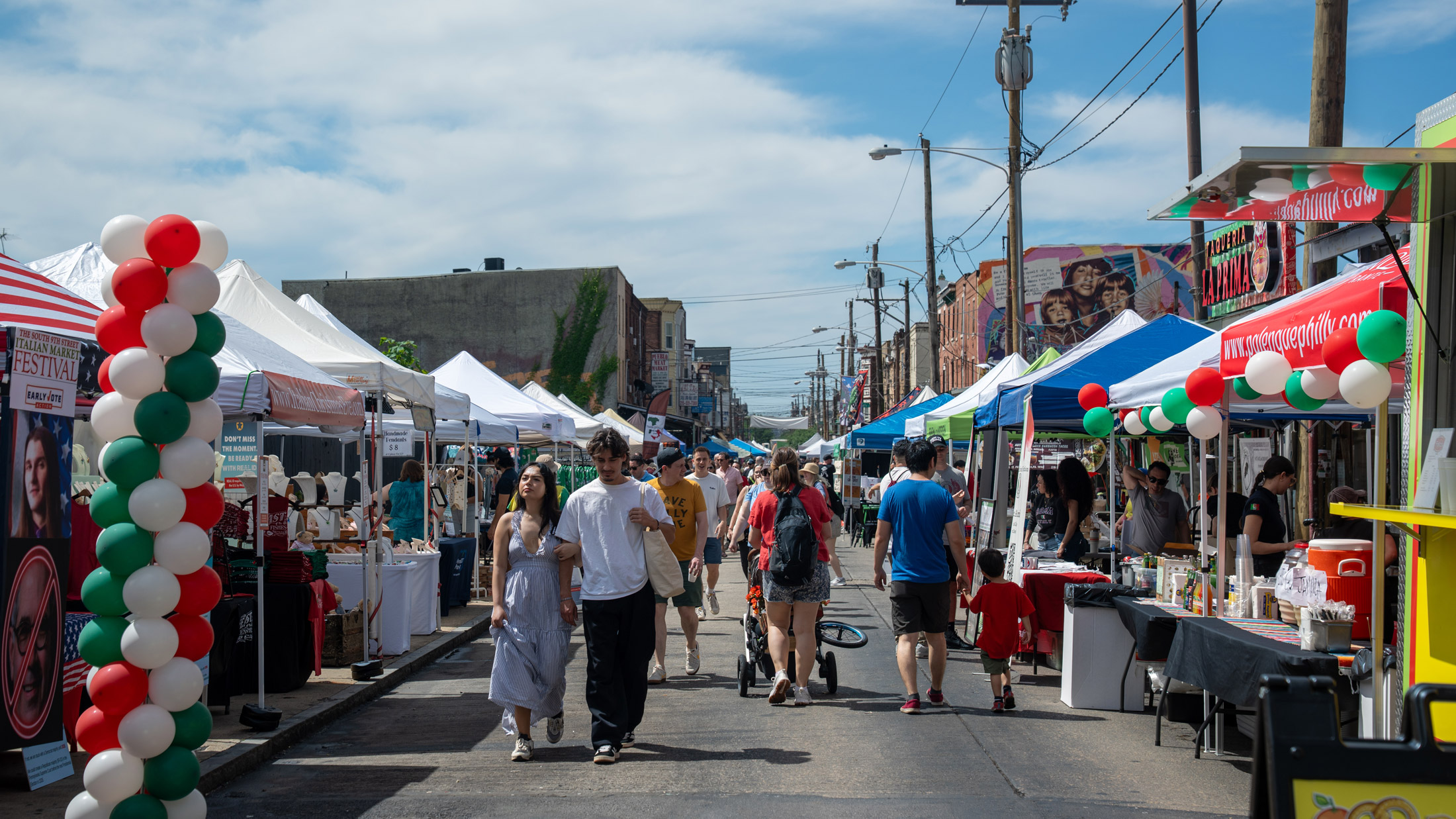 A crowded street festival scene at the Italian Market in Philadelphia, with vendor tents lining both sides of South 9th Street. The street is filled with pedestrians walking past white tents and vendor booths. Red, white, and green balloons (Italian flag colors) are clustered on the left side. Historic brick buildings line the background, and blue sky with light clouds fills the upper portion of the image. Colorful mural artwork is visible on a building to the right.