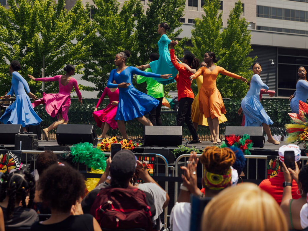 Dancers performing at Philadelphia's Juneteenth Block Party on an outdoor stage, wearing vibrant colorful costumes in shades of blue, red, yellow, and orange, with a crowd of spectators watching below and festive floral decorations and greenery surrounding the stage.