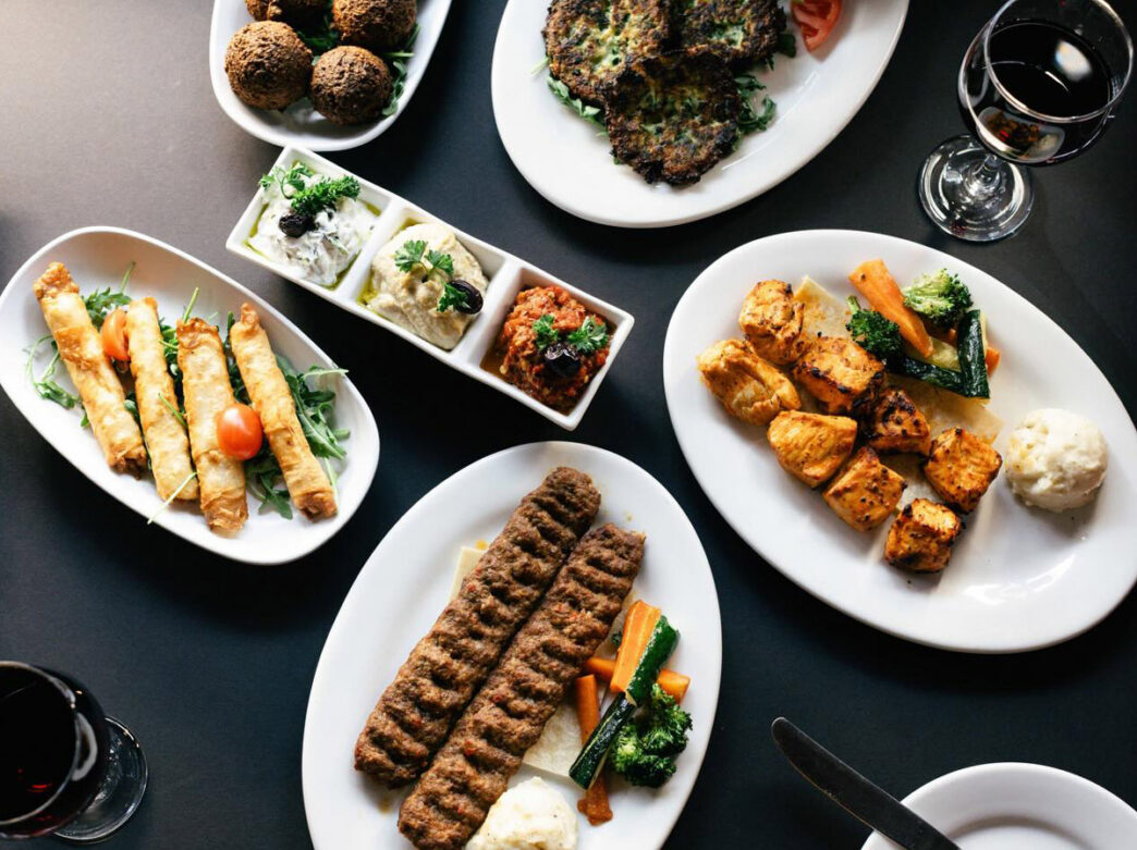An overhead view of a Mediterranean dining table featuring multiple white plates of grilled and fried dishes including falafel, kebabs, grilled vegetables, and seasoned meat, arranged around a center plate with hummus and other dips. Glasses of red wine and water are visible on the dark table surface.