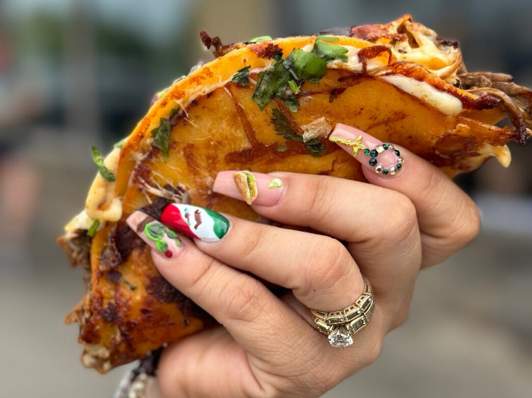 A woman with painted nails holds a crispy taco at La Catrachita Taqueria