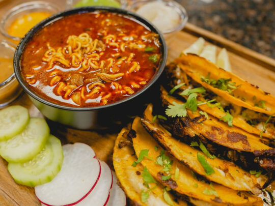 Des tacos sont posés sur une table à côté de légumes frais et d'une soupe ramen au birria à La Catrachita Taqueria.