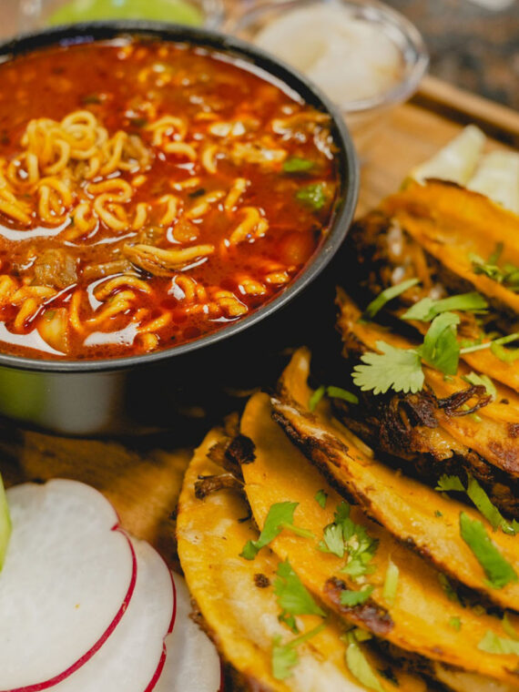 Tacos sit on a table next to fresh vegetables and a birria ramen soup at La Catrachita Taqueria