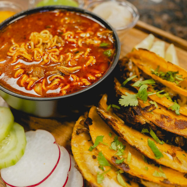 Des tacos sont posés sur une table à côté de légumes frais et d'une soupe ramen au birria à La Catrachita Taqueria.
