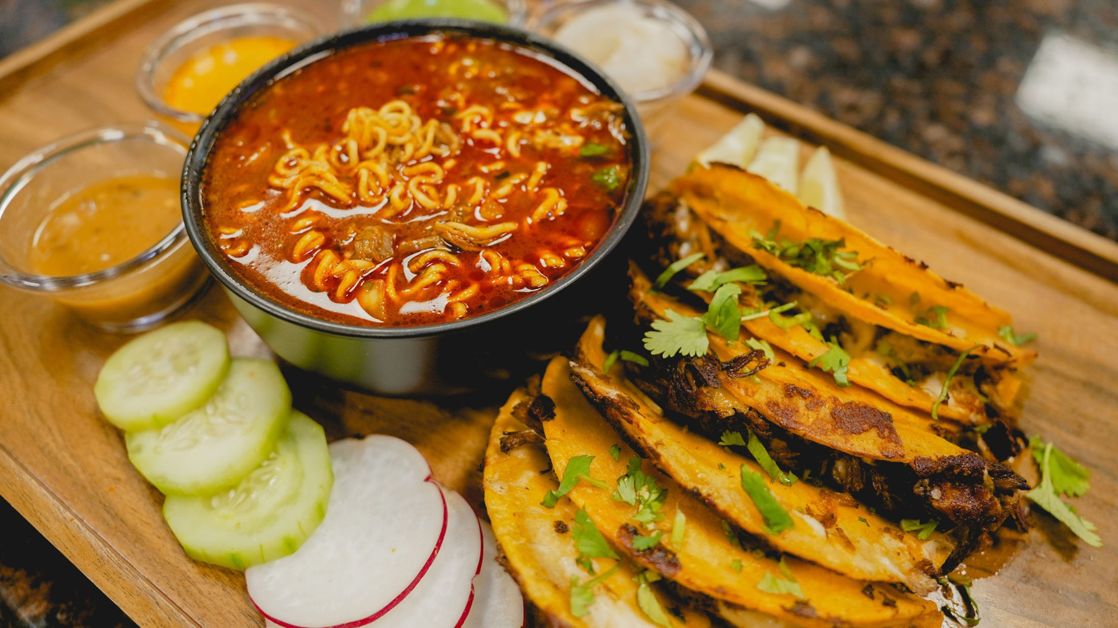 Tacos sit on a table next to fresh vegetables and a birria ramen soup at La Catrachita Taqueria