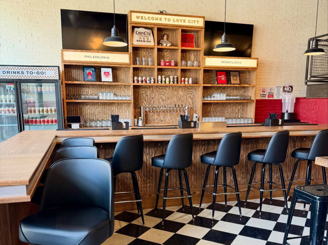L'intérieur de la brasserie Love City Brewing à Manayunk, Philadelphie, présente une salle de dégustation moderne avec un comptoir de bar en bois et des tabourets de bar noirs. Derrière le bar se trouve une grande étagère en bois portant la mention "Welcome to Love City", sur laquelle sont exposés des articles de marque, des verres et des objets de décoration. Les étagères sont flanquées de panneaux indiquant "Philadelphie" et "Pennsylvanie" et sont éclairées par des lampes suspendues de style industriel.