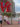 Visitors take photos at the iconic red LOVE sculpture in Philadelphia's Love Park. The large letters are stacked with blue and purple elements visible through the "O" and "E." Historic brick and modern high-rise buildings surround the plaza, with green landscaping, a fountain, and red chairs visible in the background on a partly cloudy day.