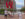 Des visiteurs prennent des photos de l'emblématique sculpture rouge LOVE dans le Love Park de Philadelphie. Les grandes lettres sont empilées et des éléments bleus et violets sont visibles à travers le "O" et le "E". Des bâtiments historiques en brique et des tours modernes entourent la place, avec un aménagement paysager vert, une fontaine et des chaises rouges visibles à l'arrière-plan par temps partiellement nuageux.