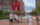 Visitors take photos at the iconic red LOVE sculpture in Philadelphia's Love Park. The large letters are stacked with blue and purple elements visible through the "O" and "E." Historic brick and modern high-rise buildings surround the plaza, with green landscaping, a fountain, and red chairs visible in the background on a partly cloudy day.