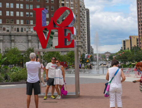 Des visiteurs prennent des photos de l'emblématique sculpture rouge LOVE dans le Love Park de Philadelphie. Les grandes lettres sont empilées et des éléments bleus et violets sont visibles à travers le "O" et le "E". Des bâtiments historiques en brique et des tours modernes entourent la place, avec un aménagement paysager vert, une fontaine et des chaises rouges visibles à l'arrière-plan par temps partiellement nuageux.