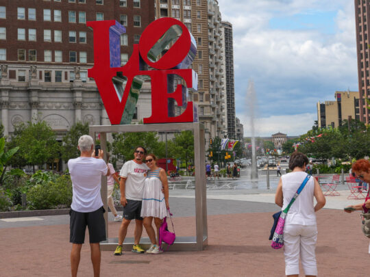 Los visitantes se toman fotos junto a la emblemática escultura roja de la palabra "LOVE" en el Love Park de Filadelfia. Las grandes letras están decoradas con elementos azules y morados que se vislumbran a través de la "O" y la «E». La plaza está rodeada de edificios históricos de ladrillo y rascacielos modernos, con zonas verdes, una fuente y sillas rojas visibles al fondo en un día parcialmente nublado.